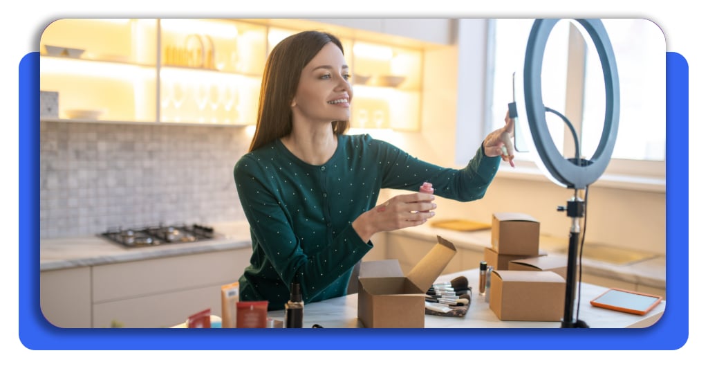 Una mujer joven está mostrando frente a su teléfono celular los productos que vende en redes sociales. Hay productos de maquillaje en la mesa.