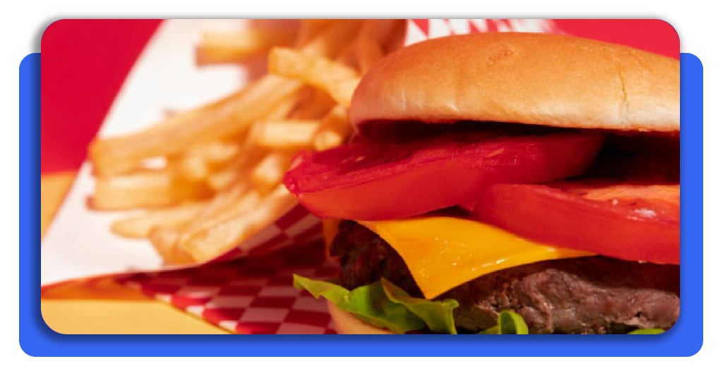 Una deliciosa hamburguesa con queso, tomate y lechuga. A su lado se observan papas fritas grandes. El pan se ve sumamente fresco.