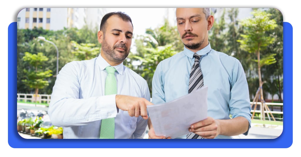 Dos hombres de negocios vestidos con camisas claras y corbata revisan un documento impreso al aire libre, detectando irregularidades fiscales.
